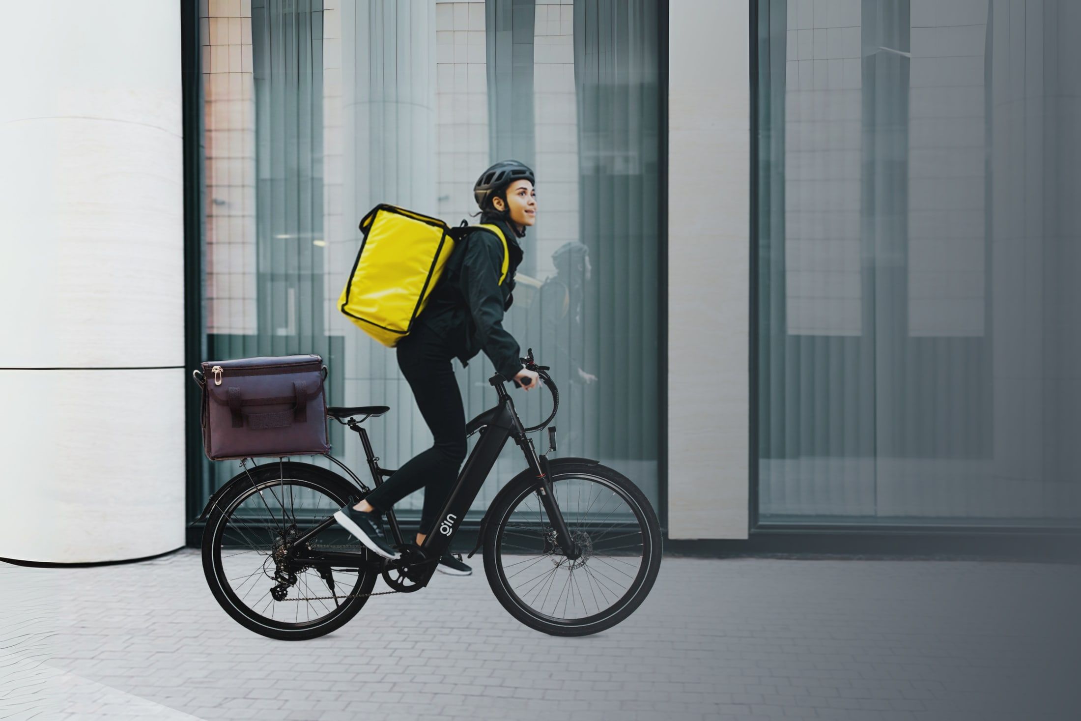 A delivery rider with a yellow bag rides a black PLUTO step-through e-bike